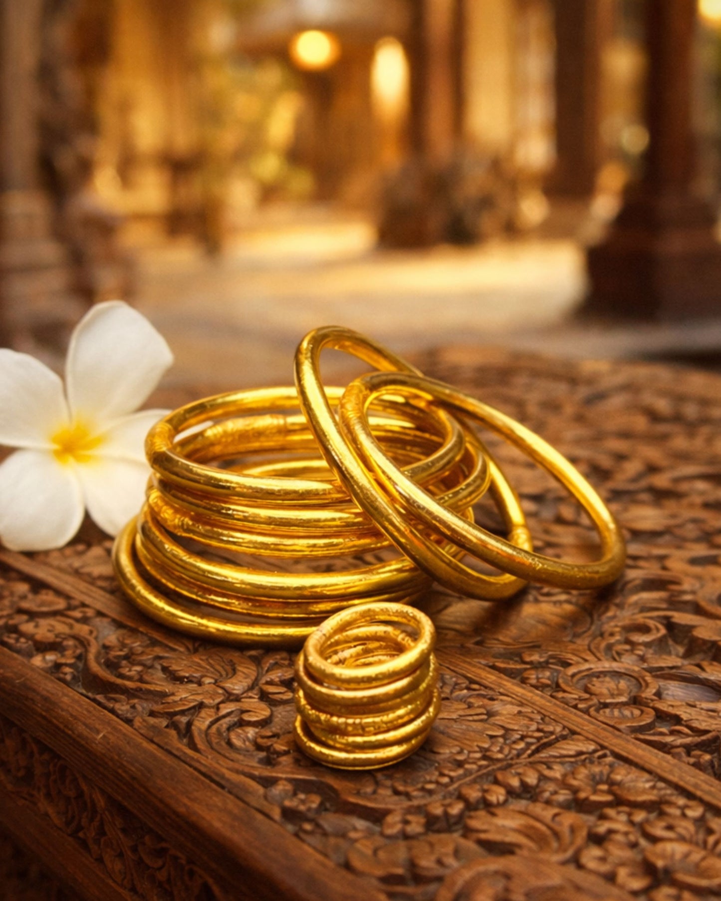 Bague bouddhiste or Bâan Kumlaï présentée avec bracelets Kumlaï dorés assortis et fleur blanche sur bois sculpté