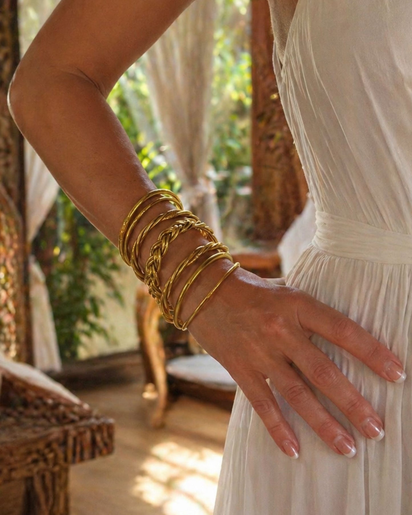 Accumulation de bracelets bouddhistes or Bâan Kumlaï avec jonc tressé, torsadé et fin, portés avec une robe blanche dans un décor chaleureux.