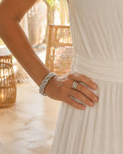 Jonc bouddhiste argent tressé Bâan Kumlaï porté seul au poignet avec robe blanche dans un décor chaleureux.