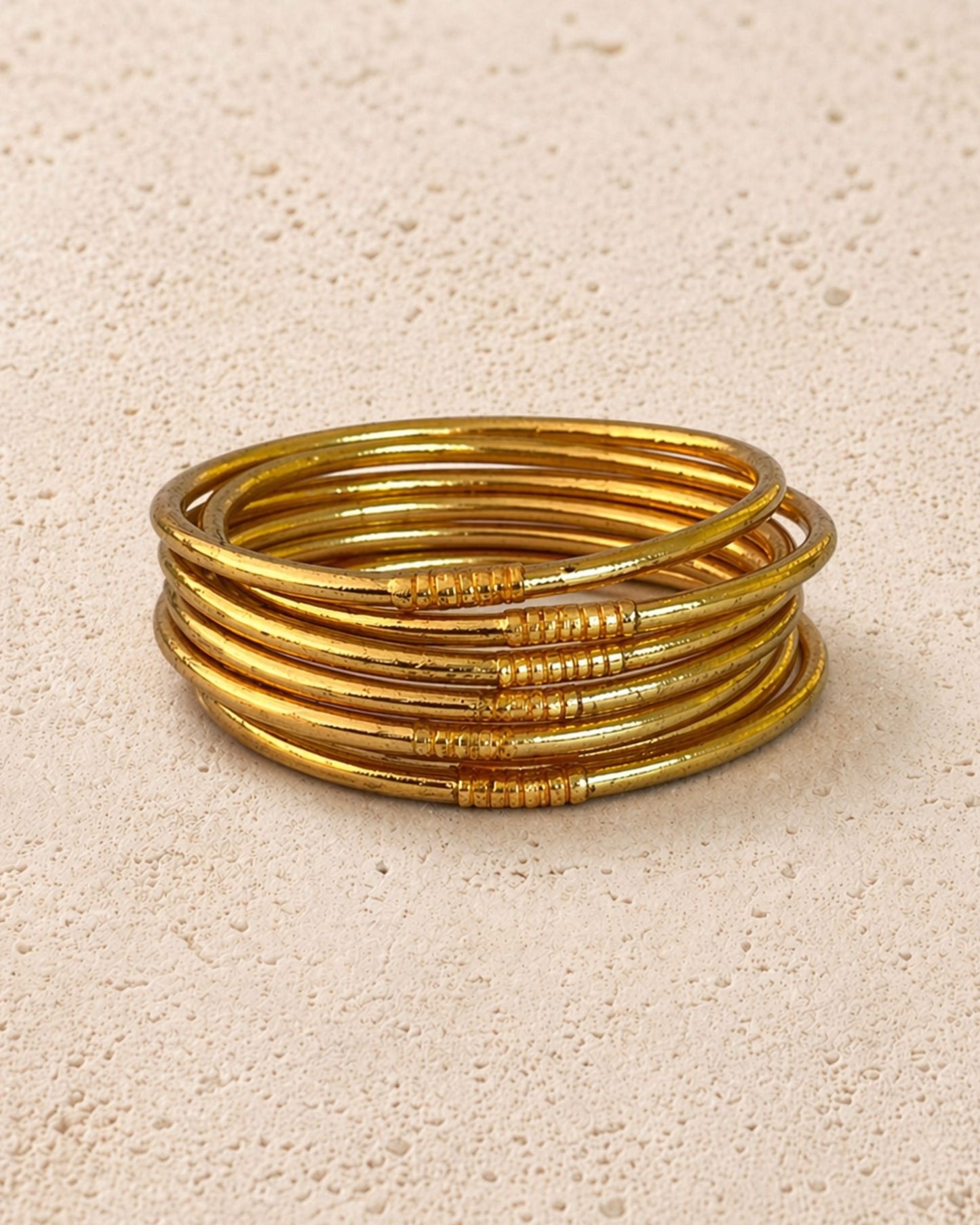 Accumulation de 7 joncs bouddhistes or fins Bâan Kumlaï sur fond minéral chaud, bracelets Kumlai dorés fabriqués en Thaïlande