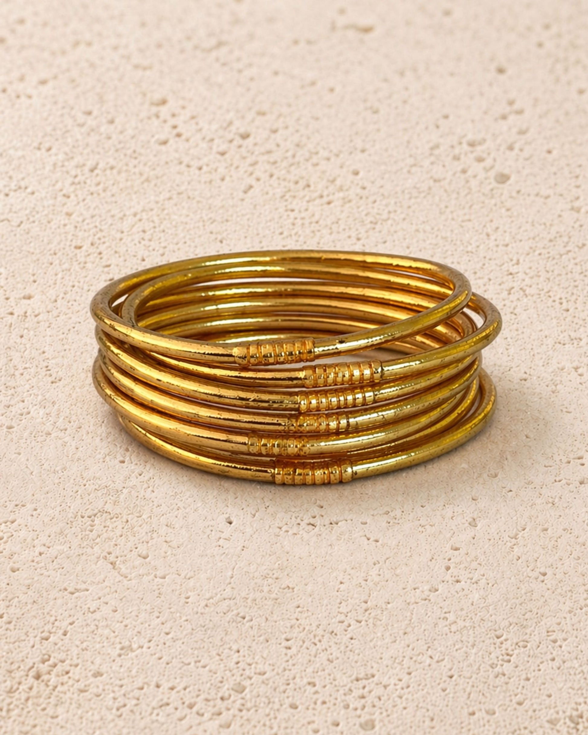 Accumulation de 7 joncs bouddhistes or fins Bâan Kumlaï sur fond minéral chaud, bracelets Kumlai dorés fabriqués en Thaïlande