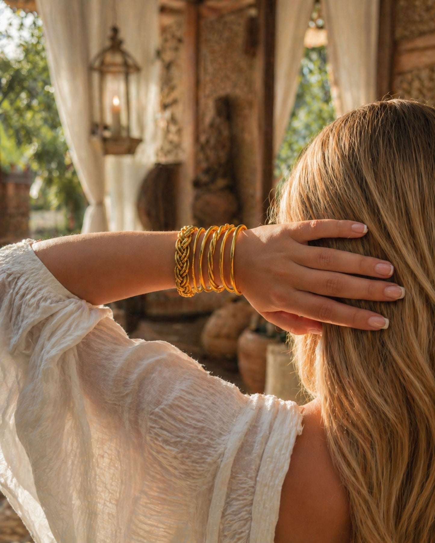 Jonc bouddhiste or tressé porté en accumulation, main dans les cheveux, avec différents bracelets Kumlai dorés assortis.