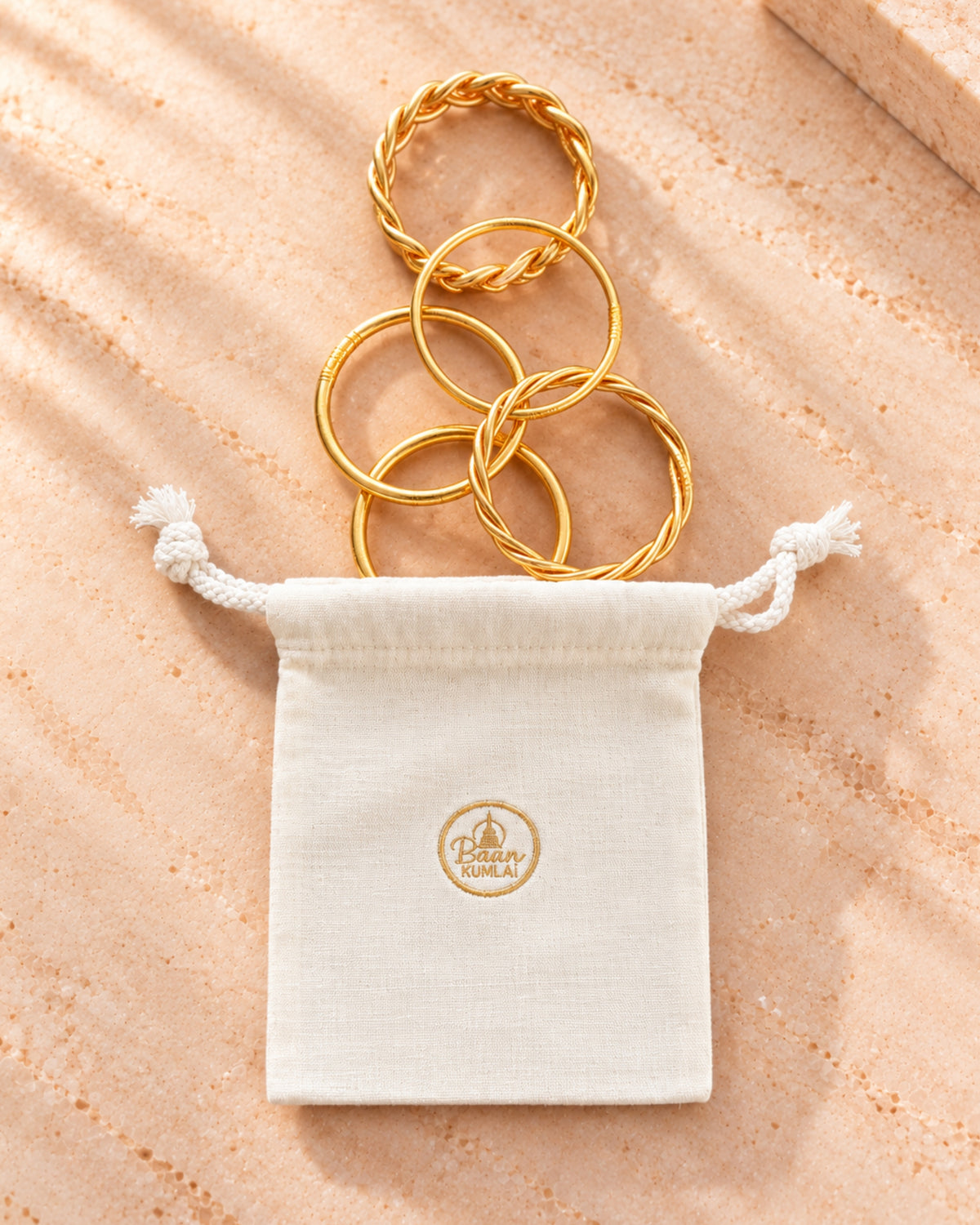 Pochon cadeau Bâan Kumlaï en tissu crème avec assortiment de bracelets bouddhistes fins, torsadés et tressés dorés.